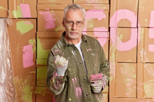 Serious grey haired handyman in glasses paints walls of room with brush busy with home renovation or apartment decoration clenches fist and looks direclty at camera wears clothes dirty with paint