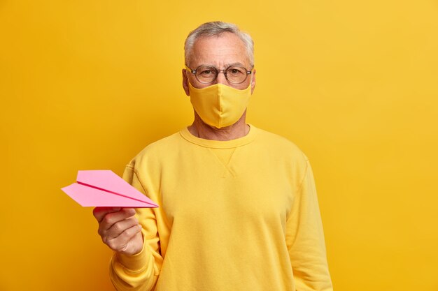 Serious grey haired man looks directly at front wears transparent glasses protective mask and holds paper airplane dressed in casual yellow jumper being infected with coronavirus