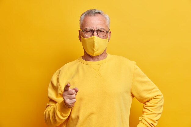 Serious grey haired man looks with strict expression at front points index finger forward weas yellow face mask as protection from virus stands indoor