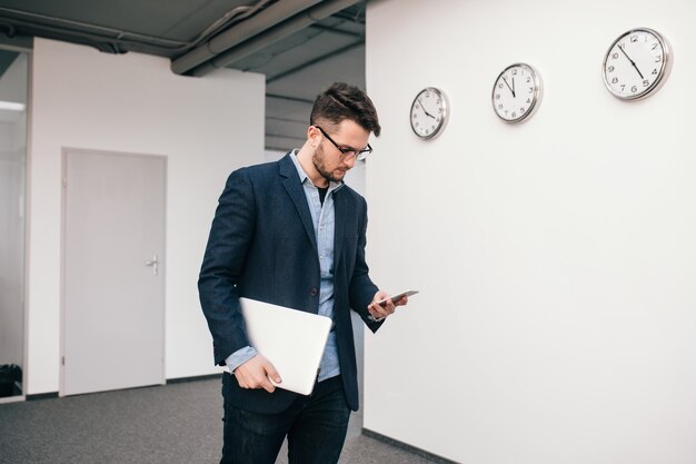 Serious guy in glasses is  walking in office. He wears blue shirt, dark jacket, jeans and beard. He is typing on phone and holds laptop in hand.