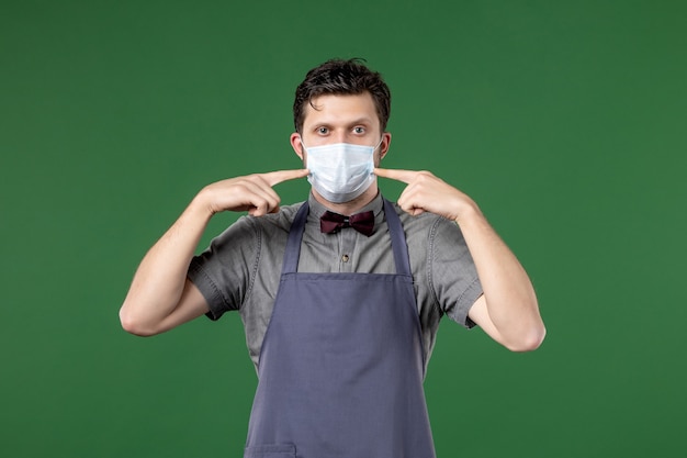 serious guy waiter in uniform with medical mask and pointing himself on green background