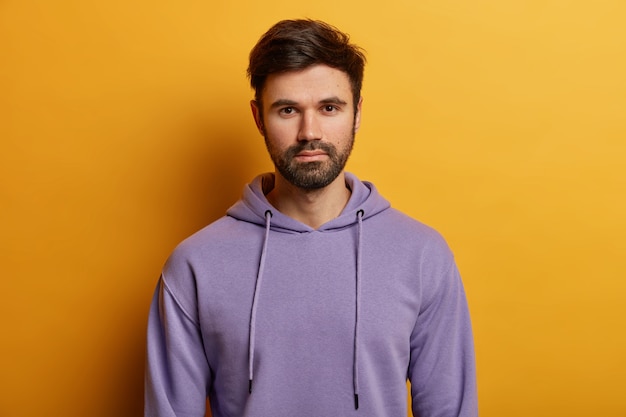 Serious handsome bearded European guy looks directly , has thick bristle, wears purple hoodie, dressed in casual hoodie, poses over yellow wall, listens information carefully.