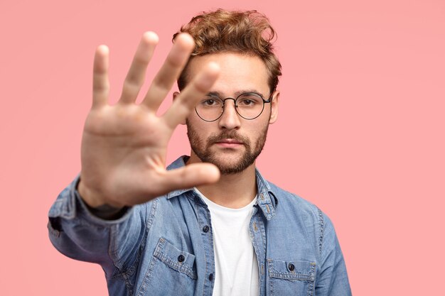 Serious handsome bearded hipster stretches hand towards camera, shows palm, demonstrates stop or hold gesture, wears fashionable clothes