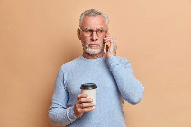 심각한 잘 생긴 수염 난 남자는 전화 대화가 귀 근처에 스마트 폰을 유지하고 커피 휴식을 즐긴다는 것은 갈색 벽 위에 고립 된 투명한 안경과 점퍼를 착용합니다.