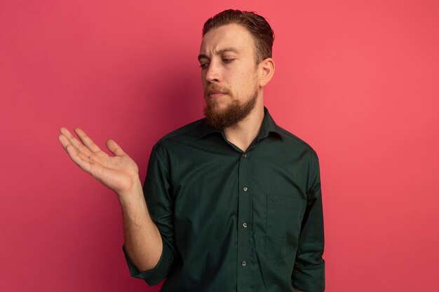 Serious handsome blonde man looks at empty hand isolated on pink wall