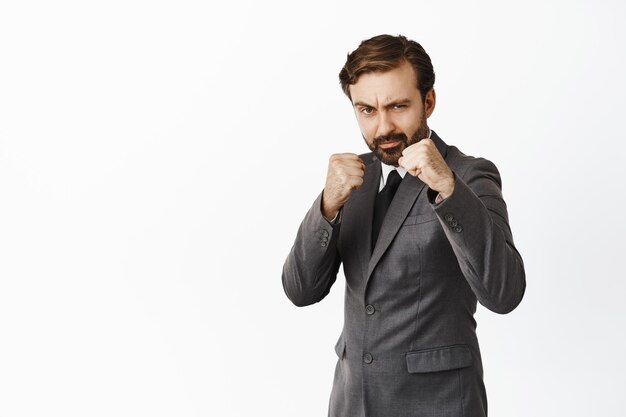 Serious handsome businessman clench fists looking at camera daring fighting boxing with someone standing over white background