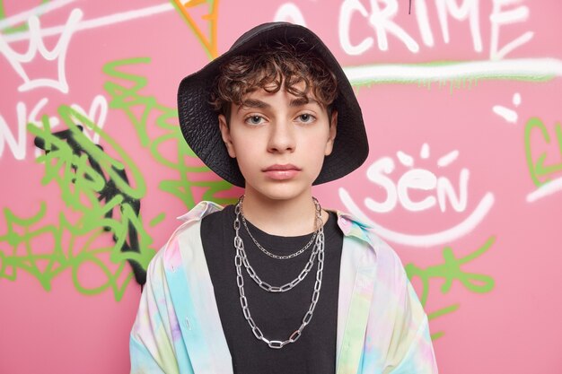 Serious handsome hipster guy looks directly at front has curly hair wears black stylish hat colorful shirt poses against colorful graffiti wall belongs to youth subculture