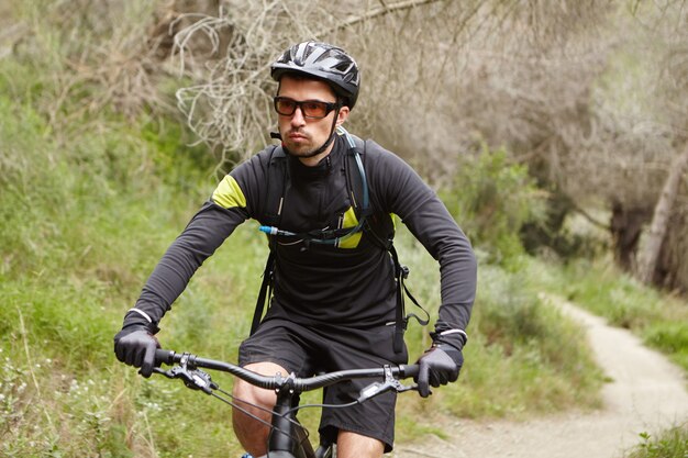 Serious handsome male biker wearing black sports clothing, helmet and eyeglasses speeding on motor-powered pedal-assist vehicle along trail in woods, having confident and self-determined look