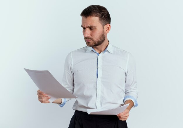 Serious handsome man holds and looks at paper sheets isolated on white wall