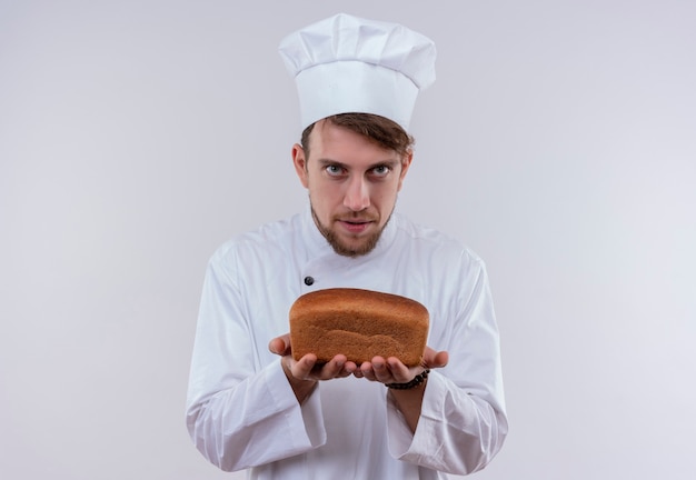 A serious handsome young bearded chef man wearing white cooker uniform and hat showing a loaf of bread while looking on a white wall