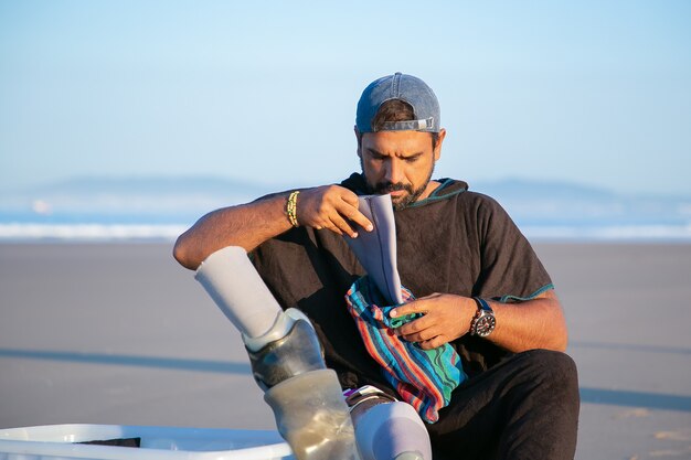 Serious handsome young man sitting on beach and putting on below-knee prosthesis