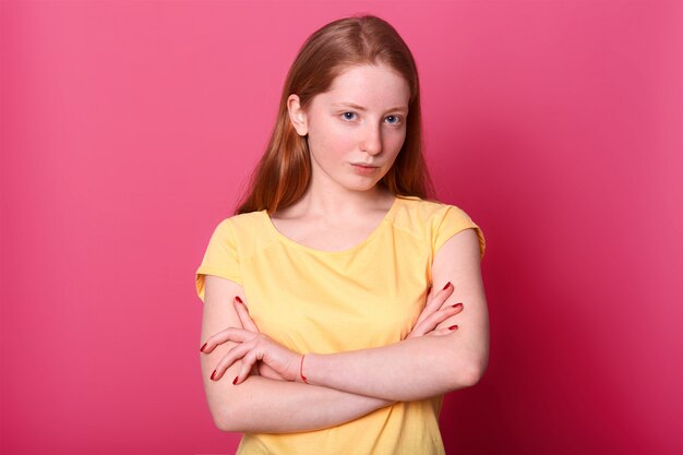 심각한 가혹한 여자 팔을 교차, 핑크에 고립 된 노란색 캐주얼 티셔츠를 입고 유지합니다. 광고 또는 판촉을위한 공간을 복사하십시오.