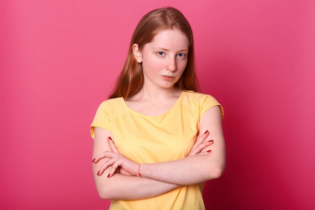 serious harsh girl keeps arms crossed, dressed yellow casual t shirt, isolated on pink. Copy space for advertisment or promotion.