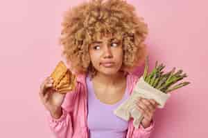 Бесплатное фото Серьезная нерешительная женщина с вьющимися волосами выбирает между вафлями и спаржей, чувствует себя сбитой с толку, одетая в повседневную куртку, сфокусированная в сторону, изолированная на розовом фоне. диета или нездоровое питание.