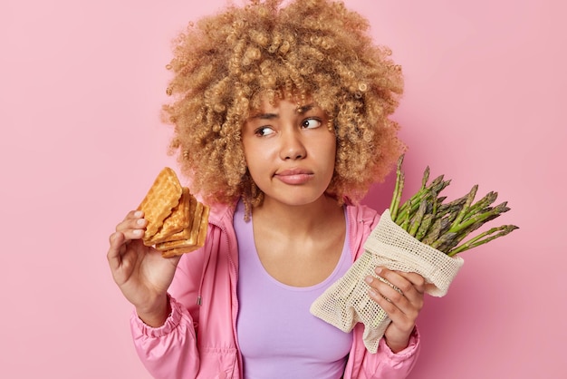 Free photo serious hesitant woman with curly hair chooses between waffles and asparagus feels confused dressed in casual jacket focused aside isolated over pink background dieting or unhealthy eating