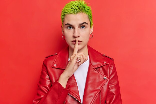 Serious hipster girl with green hairstyle makes shush gesture keeps index finger over lips zips wears leather jacket poses against vivid red wall