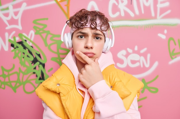 Free photo serious hipster guy holds chin tries to find solution listens favorite music via headphones dressed in fashionable clothes poses against colorful graffiti wall