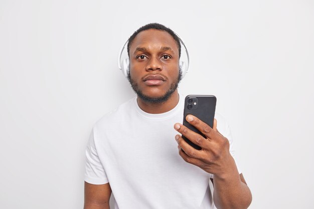 Serious hipster guy with dark skin holds mobile phone and listens to  to music via headphones