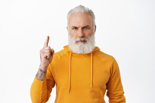 Serious hipster old man frowning pointing finger up at something important read caption showing link or advertisement above standing over white background