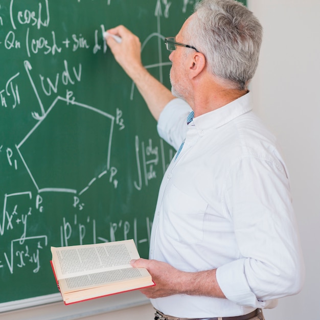 Serious lecturer in glasses chalking formula on blackboard