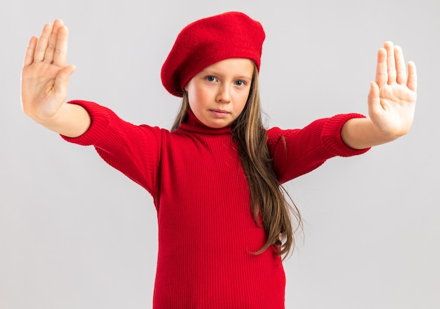 Serious little blonde girl wearing red beret showing stop gesture looking at front isolated on white wall