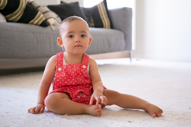 Bambina seria in pantaloncini salopette rosse seduto sul tappeto a casa. bambino a piedi nudi premuroso adorabile che si siede da solo nel salotto infanzia, fine settimana, essendo a casa concetto