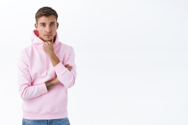 Серьезный белокурый молодой человек в розовой толстовке с капюшоном слушает скептически неинтересно