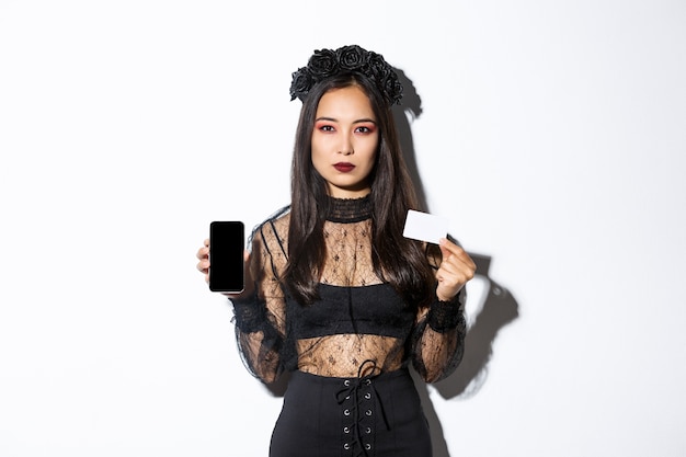 Serious-looking elegant young asian woman in gothic dress and black wreath showing credit card and mobile phone screen, celebrating halloween.