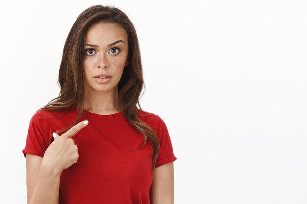 Serious-looking frustrated young woman in red t-shirt pointing herself surprised and disappointed, raise one eyebrow with disdain and disbelief, standing blank white wall intense