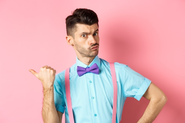 Serious-looking guy in suspenders suggessting to go outside, pointing left and looking confident at camera, starting fight, standing over pink background.