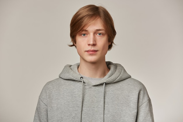 Serious looking male, handsome guy with blond hair. wearing grey hoodie. people and emotion concept. watching confident  isolated over grey wall