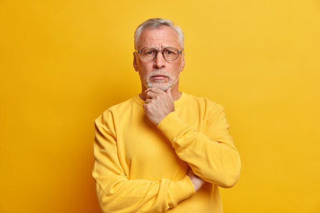 Serious looking puzzled bearded grey haired handsome man holds chin and looks directly at front dressed in casual sweater isolated over yellow wall