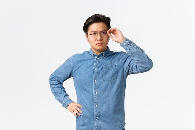 Бесплатное фото Серьезно выглядящий подозрительный азиатский предприниматель мужского пола трогает очки на лице и сомневается, стоит нерешительно или скептически, не доверяет кому-то, позирует неуверенно на белом фоне