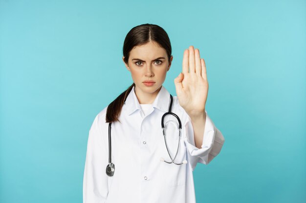 Серьезно выглядящая молодая женщина-врач, работница медицинской клиники, показывающая стоп-запрет, запрещающий жест...