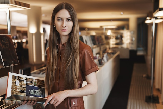 Serious-looking young female picking design magazines at vintage store, standing in stylish brown dress.