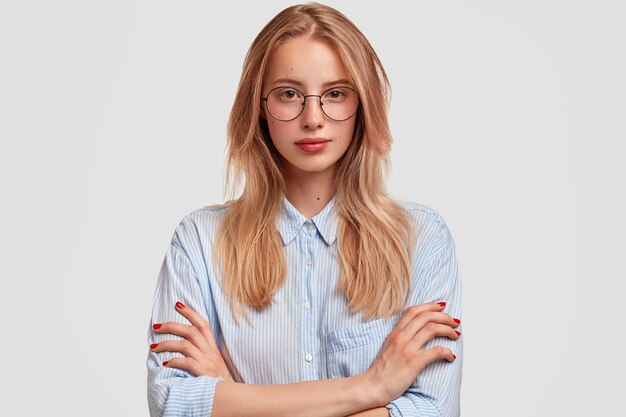 Serious lovely woman student in spectacles, keeps hands folded, listenes attentively professor`s lecture, wears elegant shirt