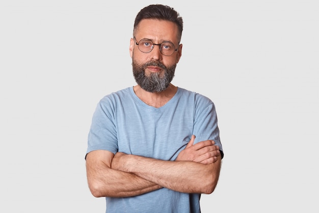 Serious magnetic black haired man posing with folded arms, having strong look, determined facial expression, having athletic arms. Middle aged bearded model poses isolated on light grey.