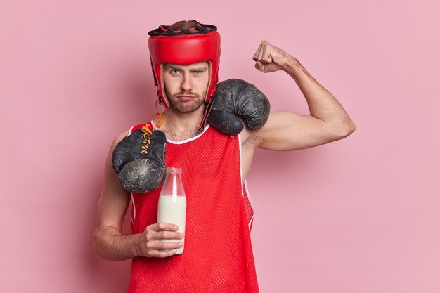 Serious male boxer raises arm shows biceps drinks fresh milk to be strong wears protective hat red t shirt boxing gloves around neck demonstrates power