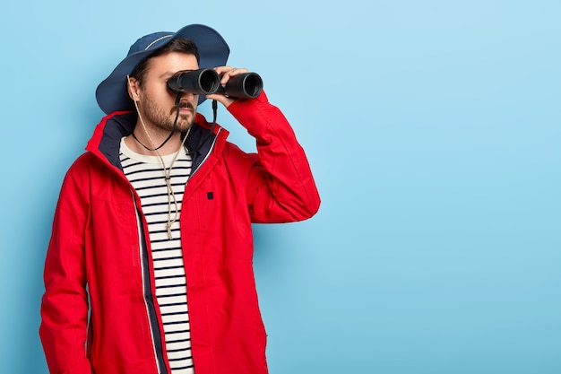 Serious male camper has long adventurous journey, keeps binoculars near eyes, wears hat and red jacket, tries to see something far away, poses against blue wall