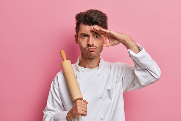 Serious male cook holds rolling pin, keeps hand on forehead, think what to cook for restaurant visitors