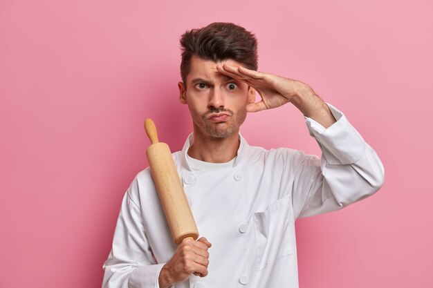 Серьезный мужчина-повар держит скалку, держит руку на лбу, думает, что приготовить посетителям ресторана