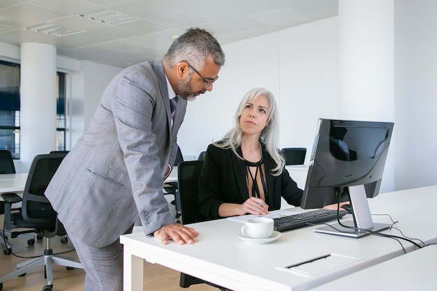 Serious male and female colleagues sitting and standing at workplace with pc, discussing paper report. Business communication concept