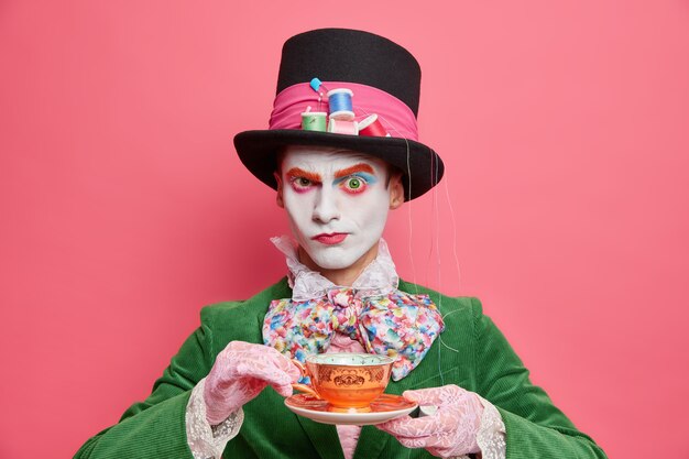 Serious male hatter from wonderland drinks tea on party looks strict at camera wears special costume ready for halloween carnival isolated over pink wall