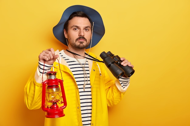 Серьезный турист-мужчина несет газовую лампу, использует бинокль во время похода, одет в плащ, уверенно смотрит в камеру, изолированную над желтой стеной