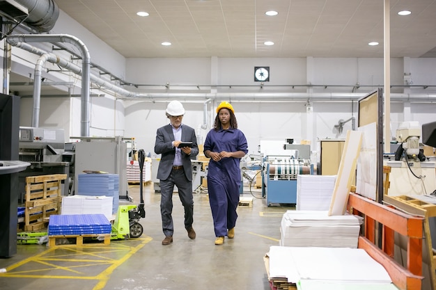 Free photo serious male inspector and female factory employee in hardhats walking on plant floor and talking, man using tablet