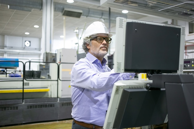 Серьезный мужчина-менеджер завода в каске и очках управляет промышленной машиной, нажимая кнопки на панели управления