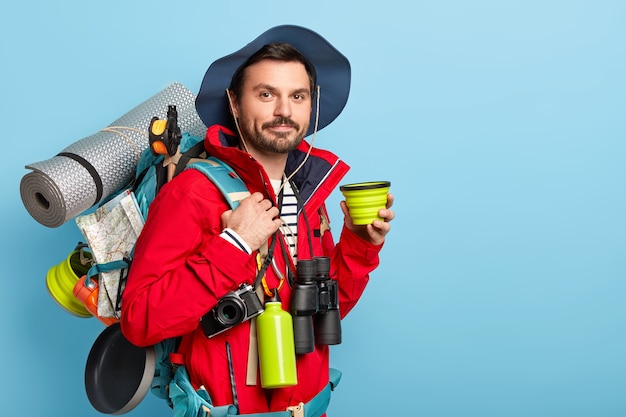 Free photo serious male tourist carries backpack with necessary equipment for traveler, enjoys traveling for long distance, prefers active vacation, drinks coffee