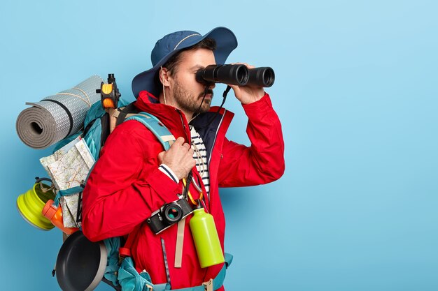 Serious male tourist uses binoculars to observe surroundings, carries rucksack with rolled up rag, map and pan for cooking on bonfire