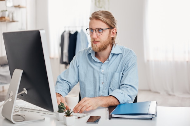 Serious male trader with blonde hair, beard, wearing glasses and blue shirt, prepares financial report on company`s income, typing on keyboard of computer, sits against modern light office interior.