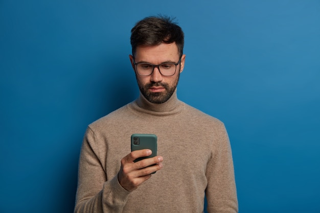 Serious male uses modern smartphone, has attentive gaze at display isolated over blue background.